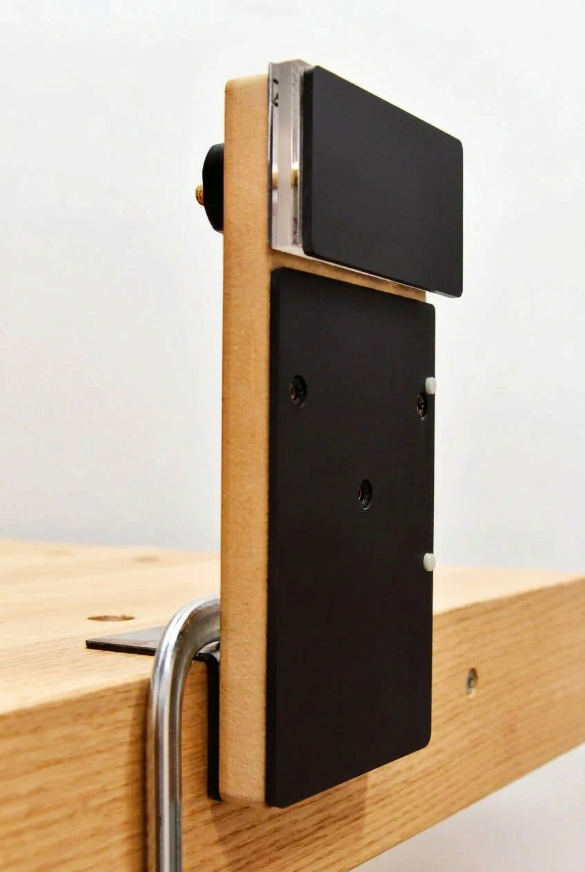 A close-up of a wooden board or frame with a black rectangular panel attached, mounted against a light gray background with part of a wooden surface and metal hardware visible.